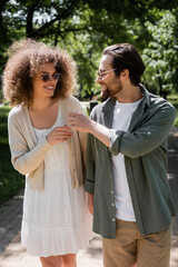curly woman and cheerful man in stylish sunglasses walking in park.