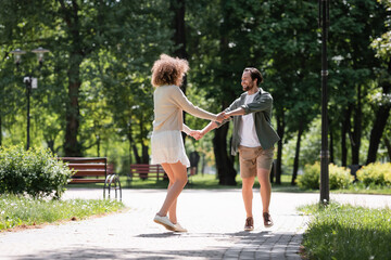 full length of joyful young couple holding hands while walking together in summer park.