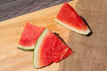 Ripe pieces of red cut watermelon with pits lie on a light yellow wooden table, against the...