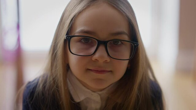 Close-up portrait of confident smiling teenage schoolgirl in eyeglasses looking at camera standing indoors. Front view headshot of charming positive Caucasian student posing in school. Education