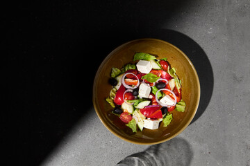 Classic greek salad on ceramic bowl in minimal style. Simple composition with greek salad on gray stone background with hard shadow. Dark concept menu. Vegetable salad on concrete table and shadow.