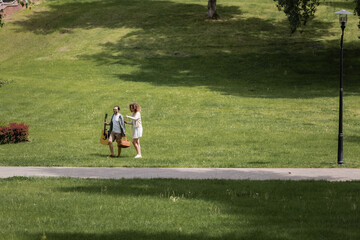 full length of couple in summer clothes carrying guitar and picnic wicker basket in green park.