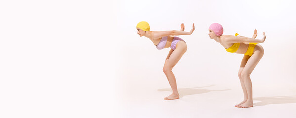 Portrait of two young women in swimming cap and suit, standing in position to dive isolated over grey studio background. Flyer