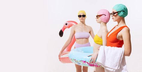 Portrait of young women in swimming suit, cap and sunglasses posing with swimming circle isolated over grey studio background. Flyer