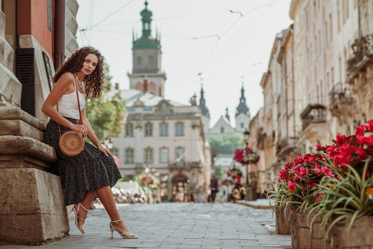 Elegant curly brunette woman wearing trendy summer outfit with round wicker shoulder bag, white top, polka dot midi skirt, strap sandals, posing in street of European city. Copy, empty space for text