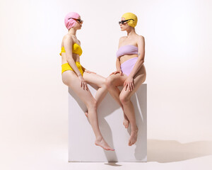 Two stylish young women in swimming suit sitting, posing isolated over grey studio background....