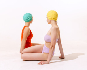 Portrait of two young women in swimming suit and cap sitting, posing isolated over grey studio...