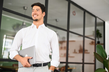 Fototapeta na wymiar High-skilled smiling young businessman carrying laptop in a modern office, white collar worker ready for new working day, inspired with new ideas male start-up owner looking aside
