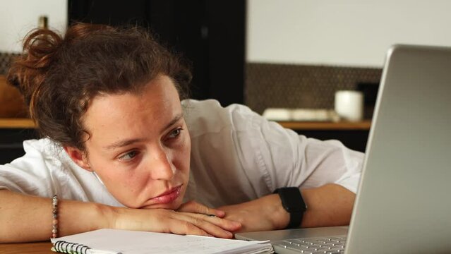 Tired young woman writing notes, using laptop for blah online education, sitting on desk at home indoor. Female student watching boring webinar,learning language, listening uninteresting audio course