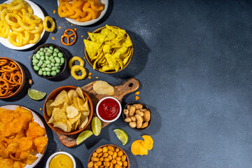 Various unhealthy salty bar snacks set. Dark grey table background with traditional party snacks - chips, onion rings, salted nuts, crisps, pretzels and sauces top view copy space