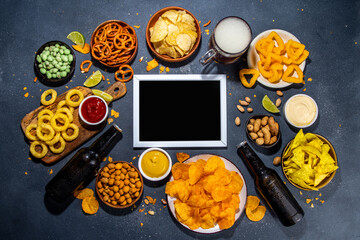 Beer with various salted snacks set. Black table background with traditional party snacks, beer bottles and glasses, with chips, onion rings, salted nuts, crisps and sauces top view copy space