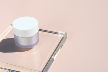 White cosmetic jar on glass stand on pink background in rays of sunlight. White glass jar for cream, lotion or mask