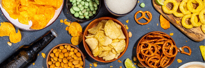 Beer with various salted snacks set. Black table background with traditional party snacks, beer bottles and glasses, with chips, onion rings, salted nuts, crisps and sauces top view copy space