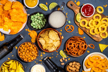 Beer with various salted snacks set. Black table background with traditional party snacks, beer bottles and glasses, with chips, onion rings, salted nuts, crisps and sauces top view copy space