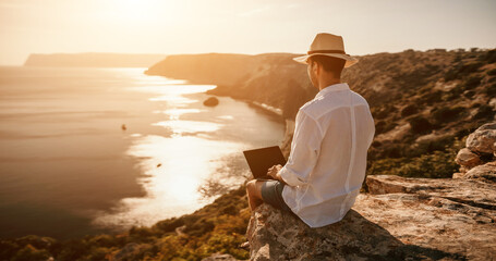 Digital nomad, freelancer. Remote work on vacation. Man in the hat, a businessman with a laptop sits on the rocks by the sea during sunset, makes a business transaction online from a distance.