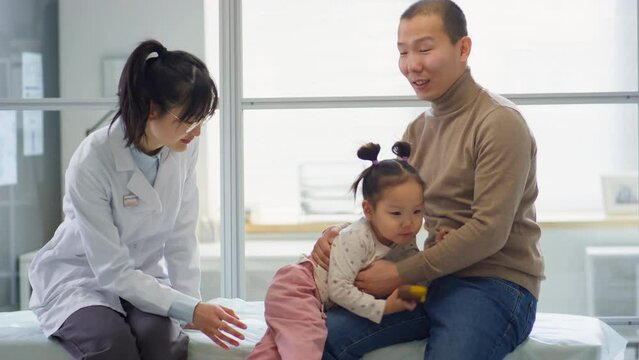 Asian father sitting with playful toddler daughter on medical couch in clinic and having consultation with female pediatrician