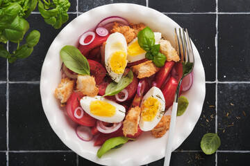 Tuscan Panzanella salad. Fresh tomatoes salad with basil leaves, eggs, olive oil and onion in white bowl on old black background. Vegetarian panzanella salad. Mediterranean healthy food. Top view.