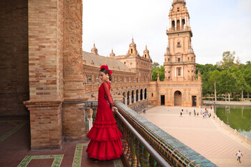 Beautiful teenage woman dancing flamenco on the balcony of a square in Seville. She wears a red...