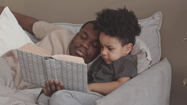 Slowmo of African American single father and his toddler son reading book while lying together in bed in evening