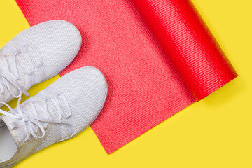 Colchoneta de yoga gimnasia roja junto a un par de zapatillas blancas sobre un fondo amarillo...