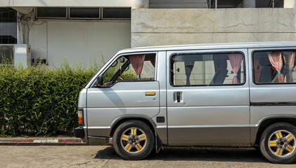A close-up view of an old gray van parked near the side of a Thai office building.