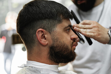 Barber guy gives a haircut to a bearded man sitting in a chair in a barbershop