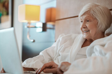 Short-haired senior woman in white robe spending time on internet and looking involved