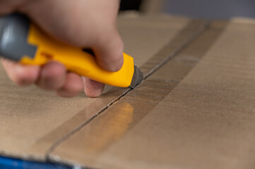 A man's hand opens a cardboard box with a yellow cutter. The flaps of the package are sealed with...