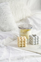 Three candles - bubble candles and white candle in glass jar with silver lid on marble tablet on white bed sheets with grey fluffy pillows