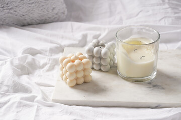 Three candles - bubble candles and white candle in glass jar with silver lid on marble tablet on white bed sheets with grey fluffy pillows