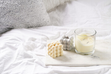 Three candles - bubble candles and white candle in glass jar with silver lid on marble tablet on white bed sheets with grey fluffy pillows