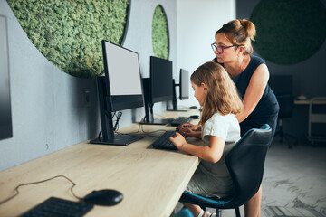 Teacher assisting schoolgirl while computer class at primary school. Child using computer on elementary computer science class. Teacher helping to find task solution. Giving clues. Back to school