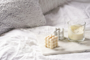 Three candles - bubble candles and white candle in glass jar with silver lid on marble tablet on white bed sheets with grey fluffy pillows