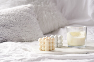 Three candles - bubble candles and white candle in glass jar with silver lid on marble tablet on white bed sheets with grey fluffy pillows
