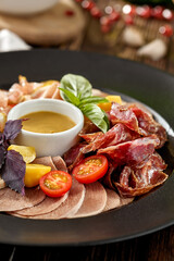 Meat platter on black plate on wooden background. Meat assorted with sauce and basil. Composition with Italian antipasti in rustic style. Meat delicatessen on dark wooden table with ingredients.