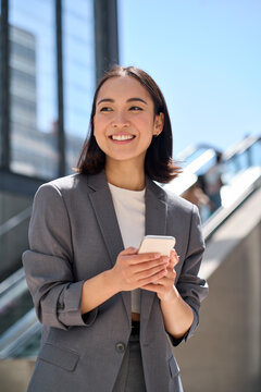 Cheerful Young Asian Business Woman Standing On Urban Street Using Cellphone. Smiling Lady Wearing Suit Holding Smart Phone Outdoor Advertising Modern City Apps, Digital Mobile Applications, Vertical.