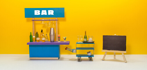 Bar service, beer drink party concept. Toy waiter trolley champagne, ice and glasses. Bar counter service area with shaker, cocktail, beer bottles spirits. Empty black chalkboard easel