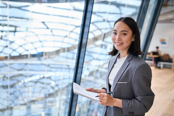 Smiling young Asian business woman manager wearing suit holding notebook standing in modern glass...