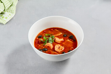 Fish tomato soup with shrimps and black olives. Seafood soup with prawns in white ceramic bowl on concrete background. Fish dish with seafood. Tomato soup in minimal composition.