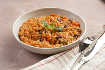Grain dish - Millet with tomatoes, beans, carrots, bell pepper and spices, mint in a white bowl on a checkered linen kitchen towel on purple background