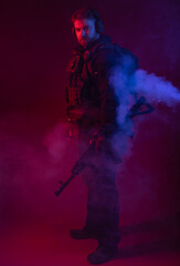 soldier in full gear with weapons. a man in headphones, body armor, with a backpack and a belt. red background. colored, blue-red light. smoke around the military. explosion, chemical attack