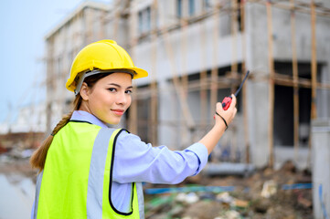 Young attractive construction woman in safety vest with yellow helmet working with radio, standing on building construction site. Home building project.