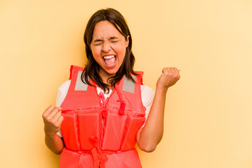 Young hispanic woman holding life jacket isolated on yellow background raising fist after a...