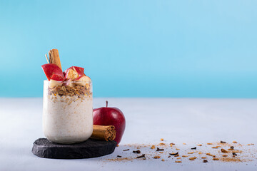 Overnight oats with apple fruits, accompanied with a whole apple and cinnamon on the back. Oatmeal, cinnamon powder, and cloves scattered. A frontal photo with a blue background for adding text.