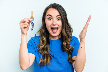 Young caucasian woman holding home keys isolated on blue background receiving a pleasant surprise,...