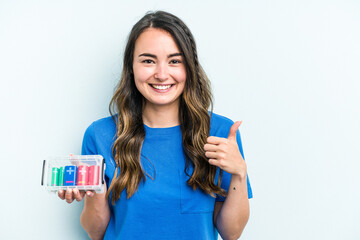 Young caucasian woman holding batteries isolated on blue background smiling and raising thumb up