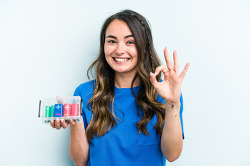 Young caucasian woman holding batteries isolated on blue background cheerful and confident showing ok gesture.