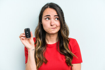 Young caucasian woman holding car keys isolated on blue background confused, feels doubtful and unsure.