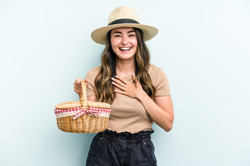 Young caucasian woman holding a picnic basket isolated on blue background laughs out loudly keeping hand on chest.