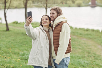 Happy young couple embracing and smiling at camera while making selfie portrait on mobile phone standing outdoors